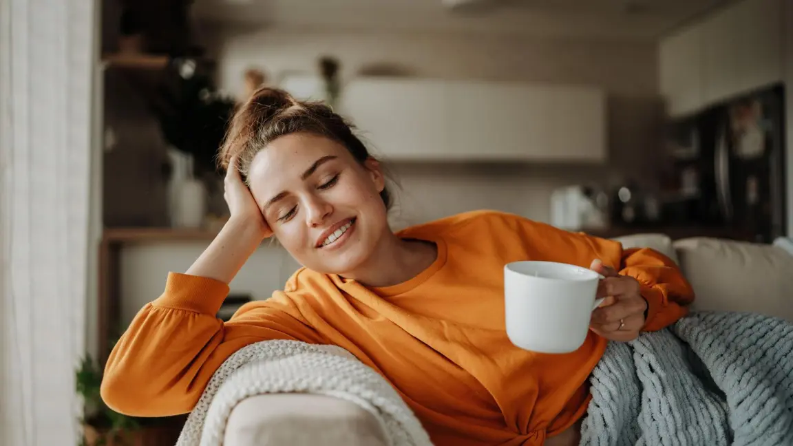 Femme souriante vêtue d’un chandail orange, assise confortablement sur un canapé, tenant une tasse blanche et semblant détendue les yeux fermés.